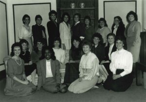 In a black and white photo, nineteen women are posing for a photo in three rows, with the back row standing and the front two rows sitting. They are dressed in 1980s era business or church attire.