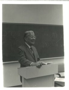 A black and white photo shows an older Eastern Asian man standing at a podium in front of a chalkboard and smiling